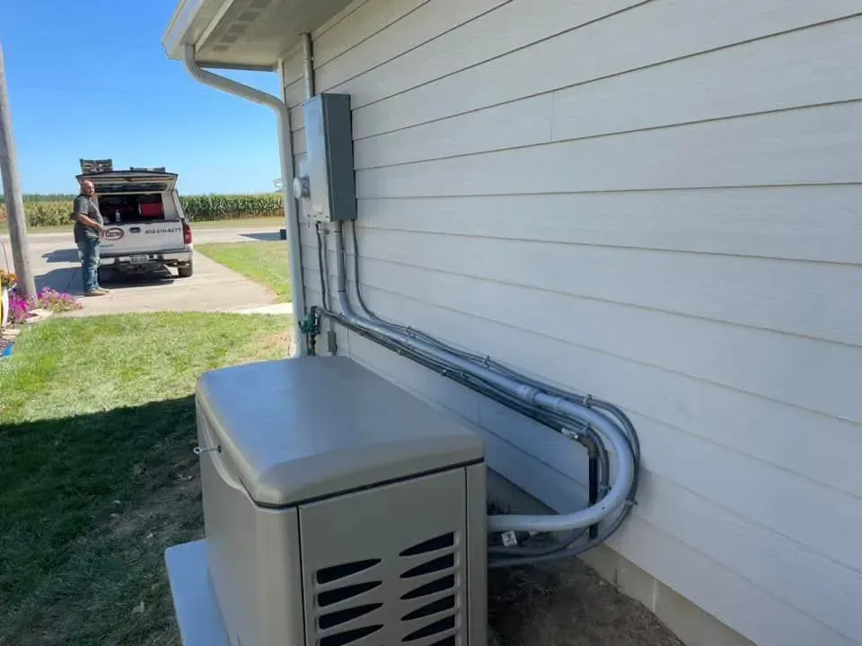 A beige standby generator sits against a house siding, connected by conduits to a gray electrical box on the wall.