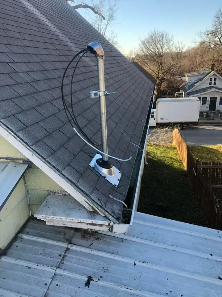 A metal mast with electrical cables mounted on a sloped shingled roof next to a metal roof section.