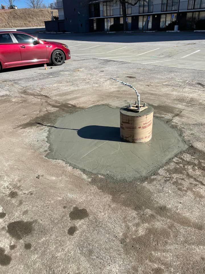 A freshly poured concrete base with a cardboard tube form stands in an asphalt parking lot near a parked red car.