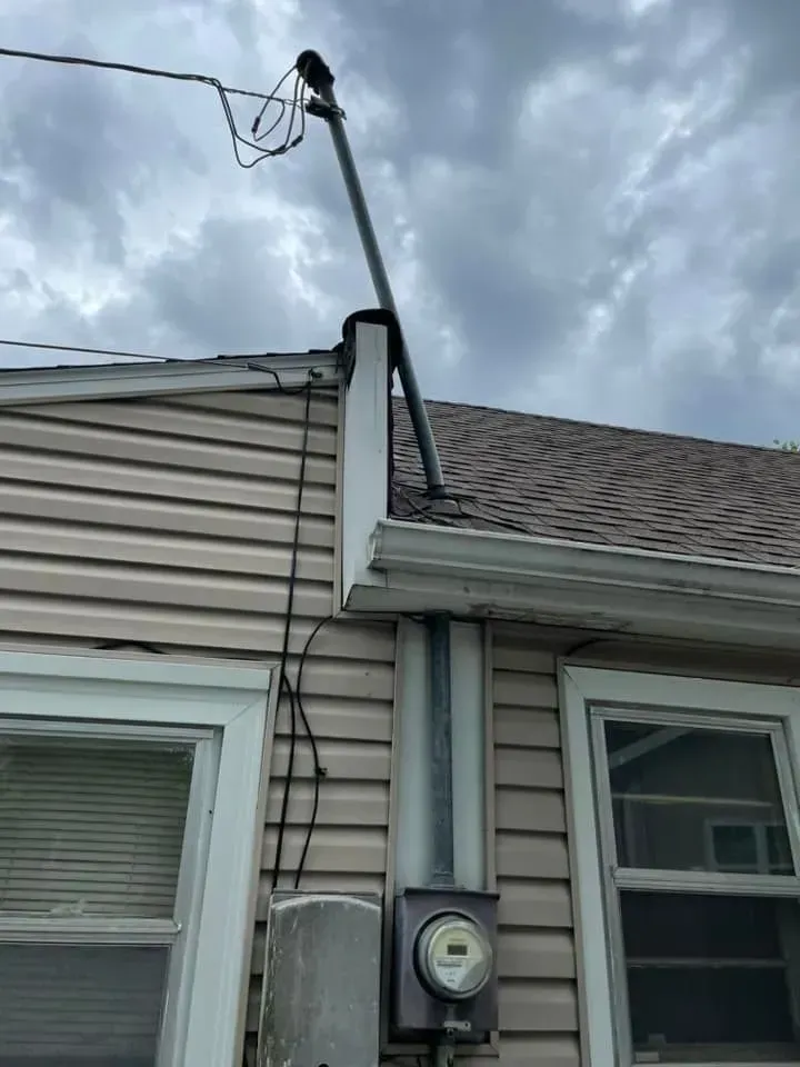 An electrical service mast rises from a roof to a utility line above a house exterior with a mounted meter box.