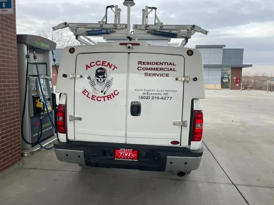 White Accent Electric service truck parked at a gas pump, showing company logo, contact info, and ladder rack on top.