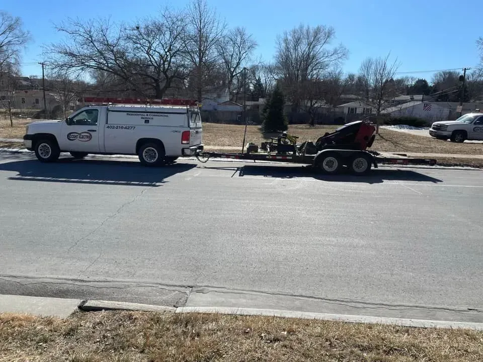 A white utility truck towing a trailer with a red piece of machinery parked on a residential street.