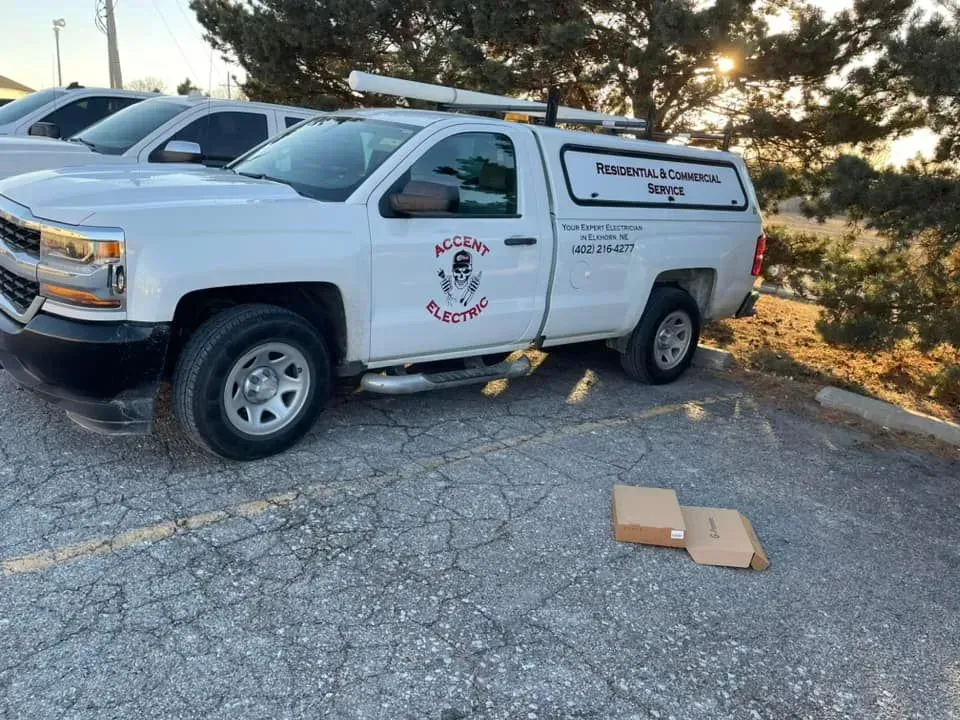 A white Accent Electric work truck parked in an outdoor lot with two small boxes on the ground next to it.