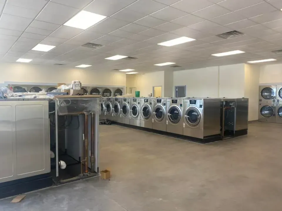 Rows of stainless-steel industrial washing machines line a brightly lit, spacious commercial laundromat.