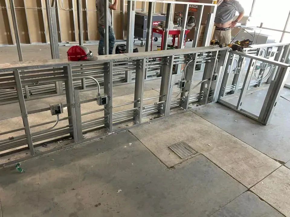 Construction site featuring metal wall framing with electrical conduits, junction boxes, and two workers in the background.