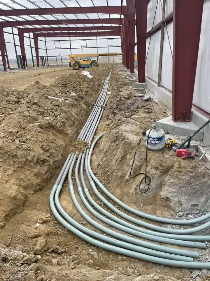 A trench inside a steel-framed building construction site contains several gray electrical conduits curving along the path.