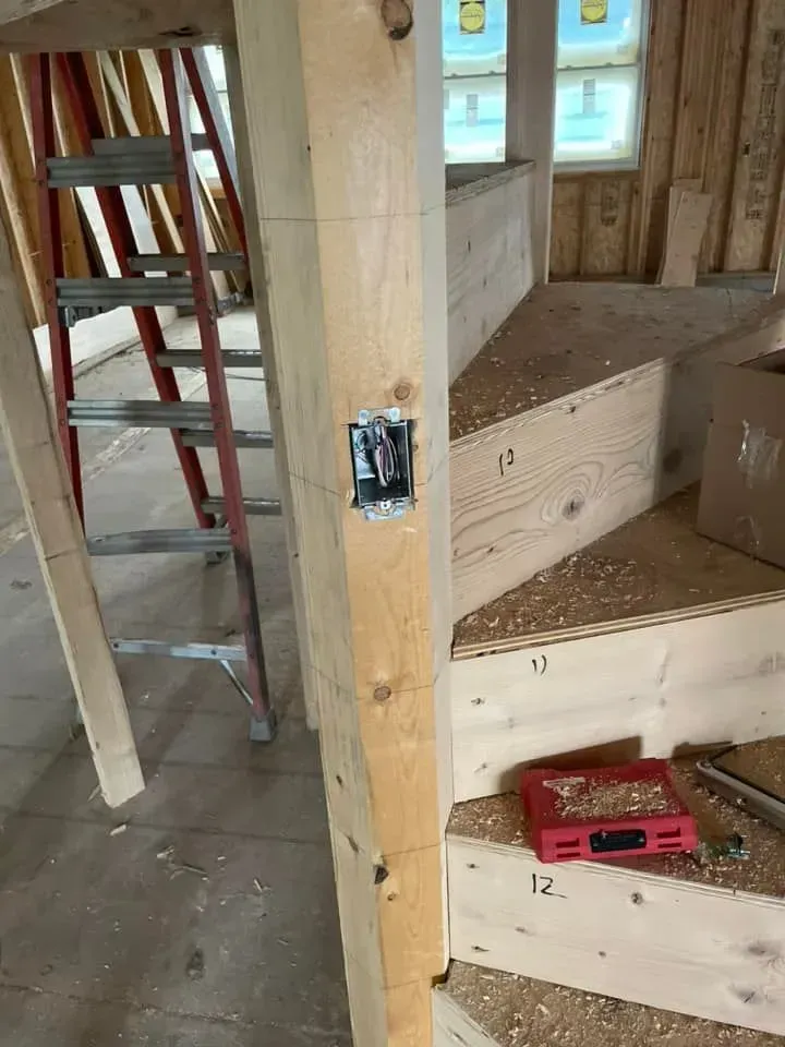 Construction site interior showing a wooden staircase under assembly and an electrical box mounted on a support post.