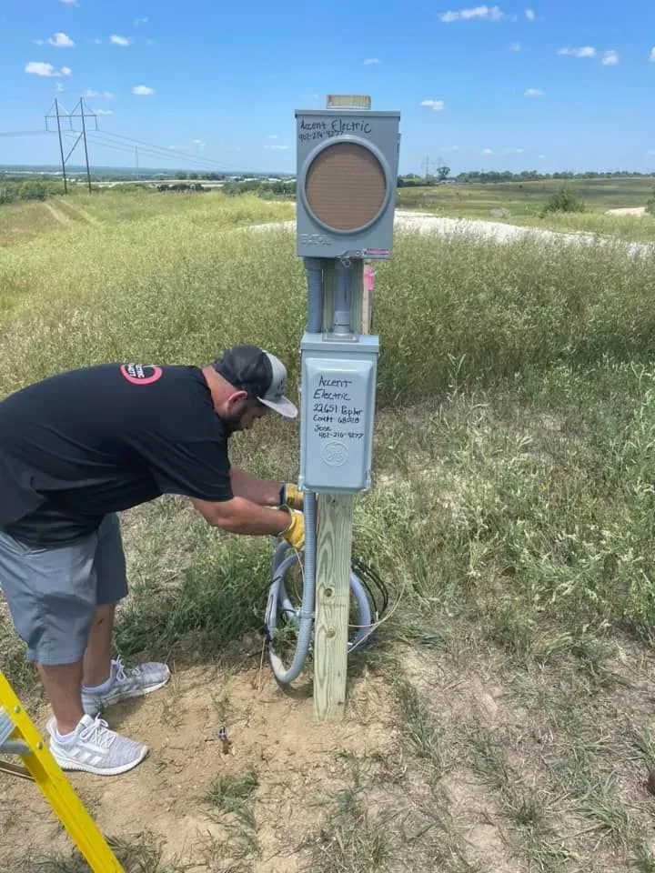 A technician in a cap and t-shirt works on an electrical service box mounted on a wooden post in an open field.