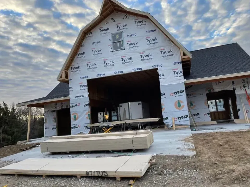A building under construction featuring a gambit roof, Tyvek house wrap, and stacks of lumber on the concrete driveway.