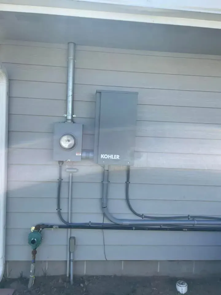 A grey Kohler transfer switch mounted on a house exterior next to an electrical meter and conduit pipes.