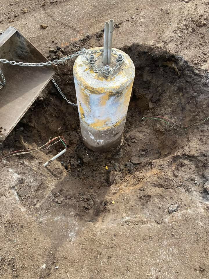 A concrete bollard stands in a dirt pit, wrapped in a chain connected to a nearby piece of machinery.