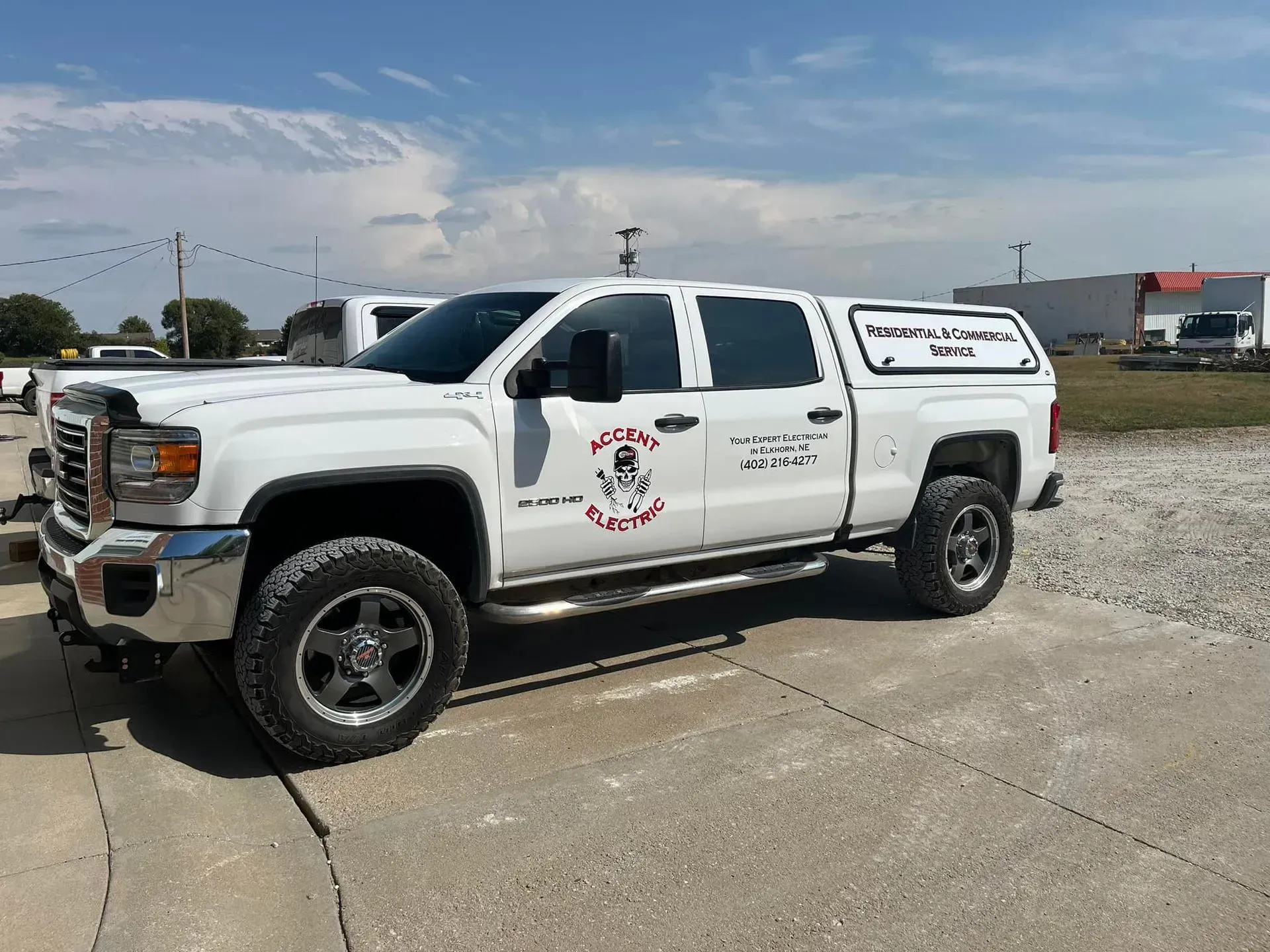 A white pickup truck with a camper shell parked on a gravel lot under a sunny sky. The side features business decals.