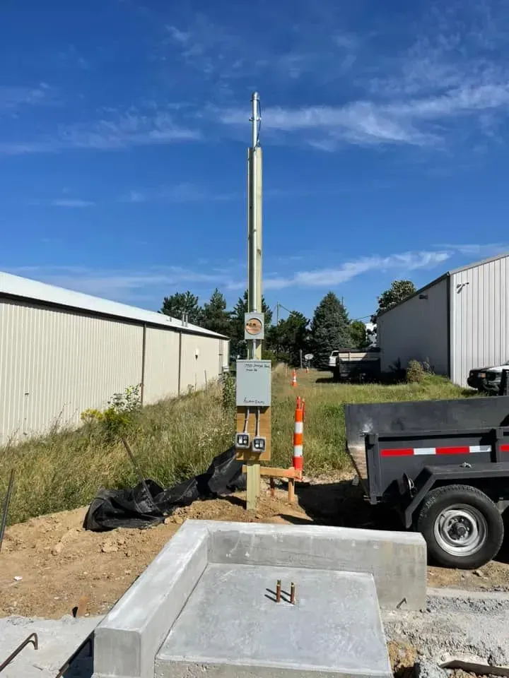 A utility pole with electrical equipment stands at a construction site next to a concrete pad and a utility trailer.
