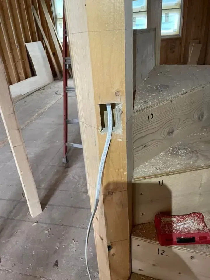 A white electrical cable hangs from a rectangular hole cut into a wooden post near unfinished stairs at a construction site.