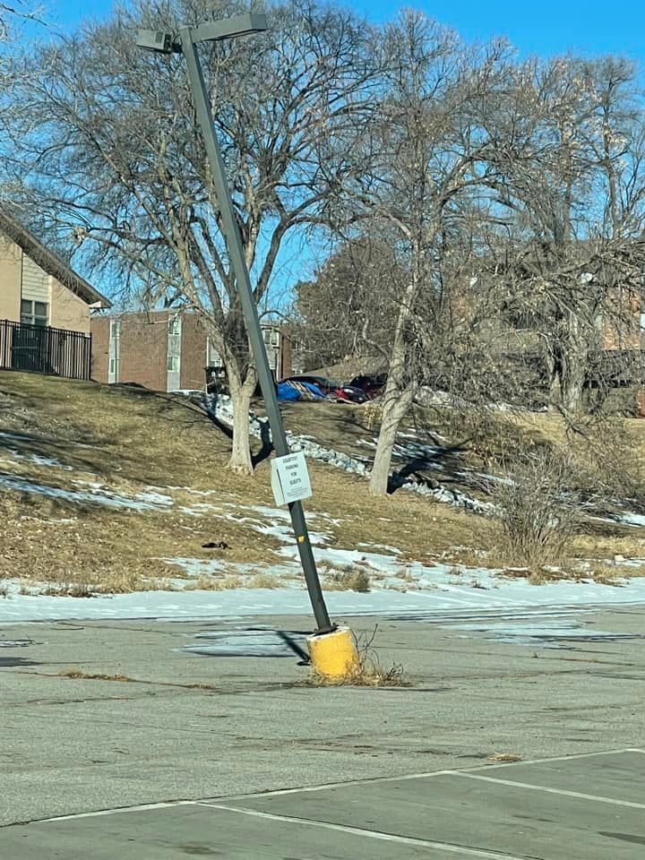A tilted metal light pole with a small sign stands in a cracked, partially snow-covered asphalt parking lot.