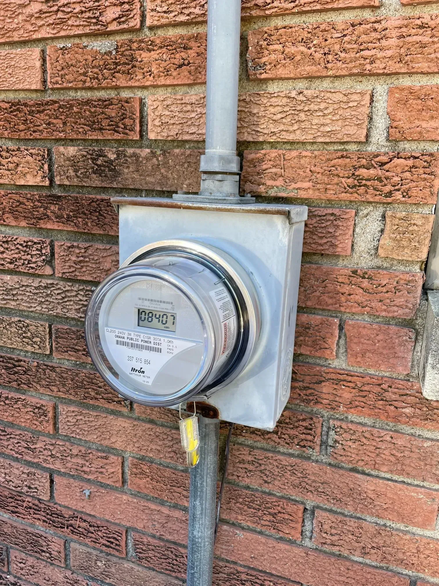 A digital electric meter mounted on a red brick wall, with a grey conduit pipe extending vertically from the top and bottom.