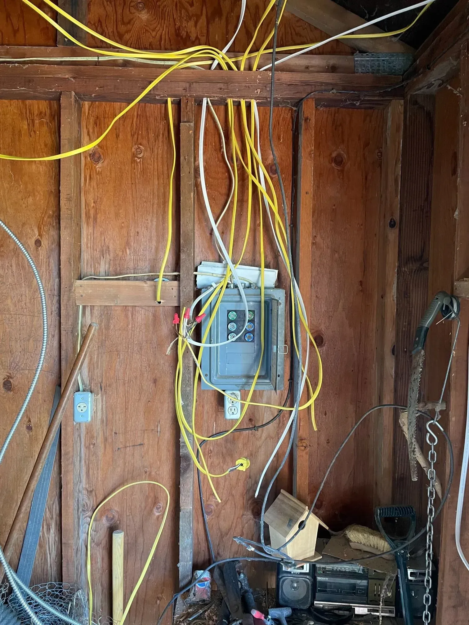 Electrical wires hang loosely from exposed wooden wall studs above a gray metal electrical breaker box in a shed.