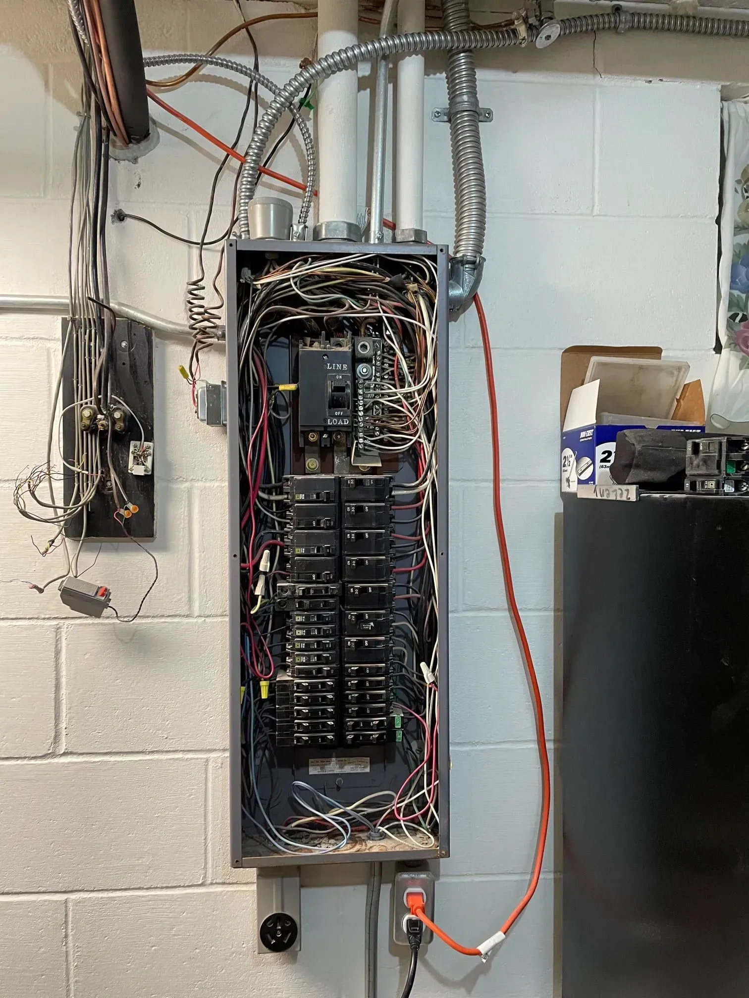 An electrical breaker panel with exposed wiring, mounted on a white cinder block wall in an unfinished basement.