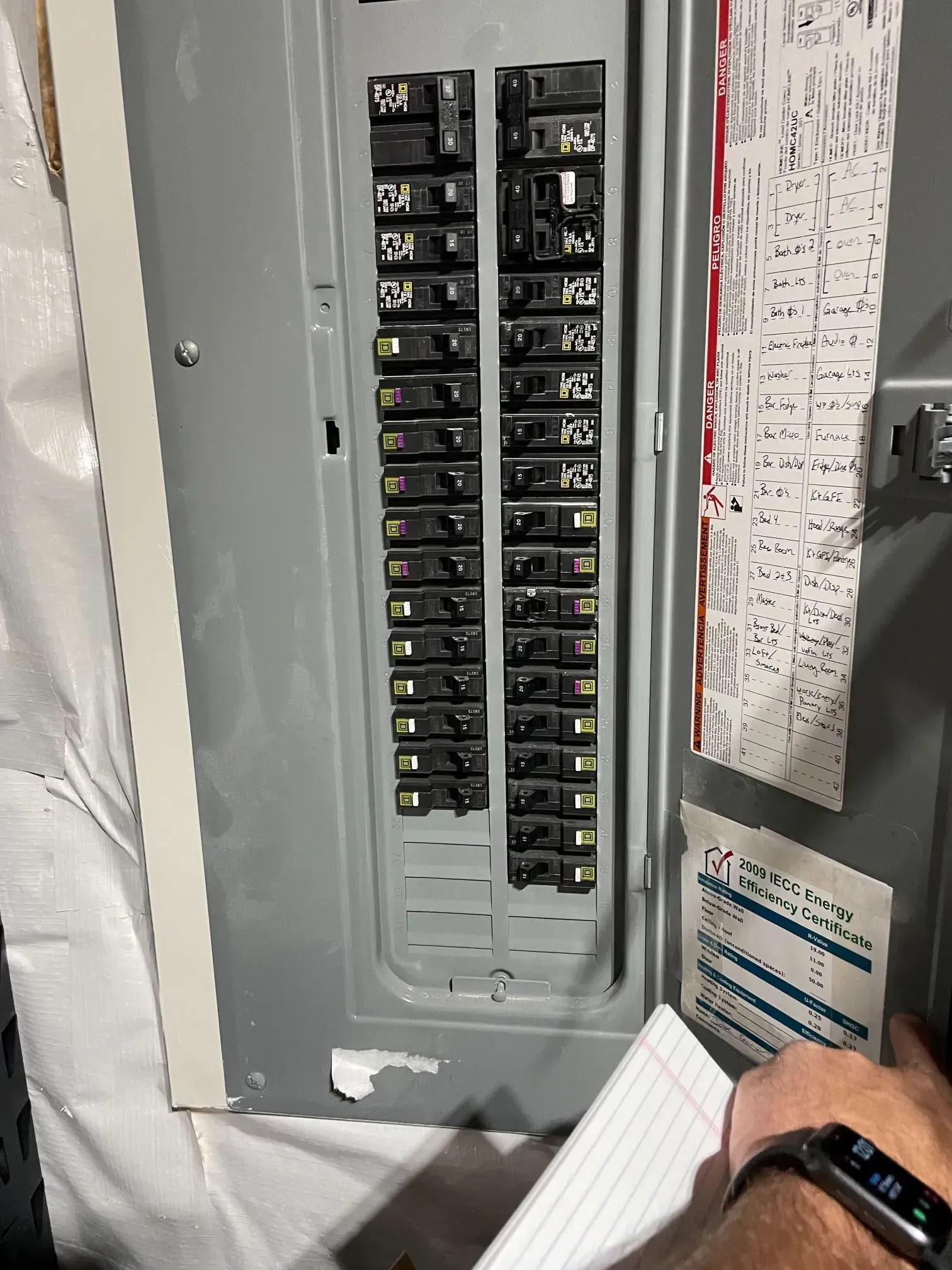 A person's hand holding a notebook points to a residential electrical circuit breaker panel with numerous switches.