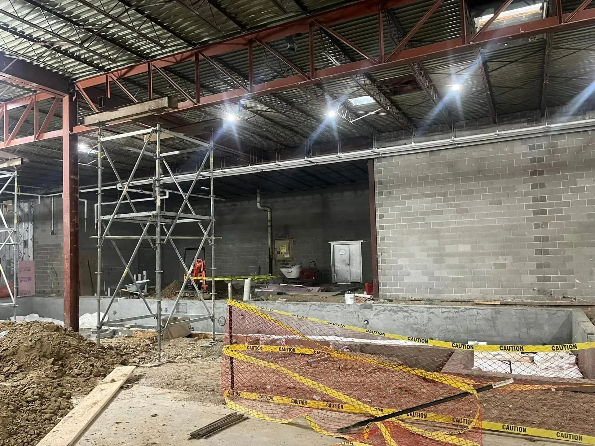 Construction site interior with scaffolding, an exposed metal ceiling, concrete block walls, and yellow warning tape.