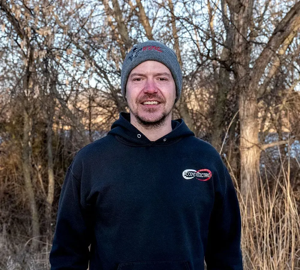 A smiling person wearing a grey beanie and a black hooded sweatshirt stands in front of bare trees at sunset.
