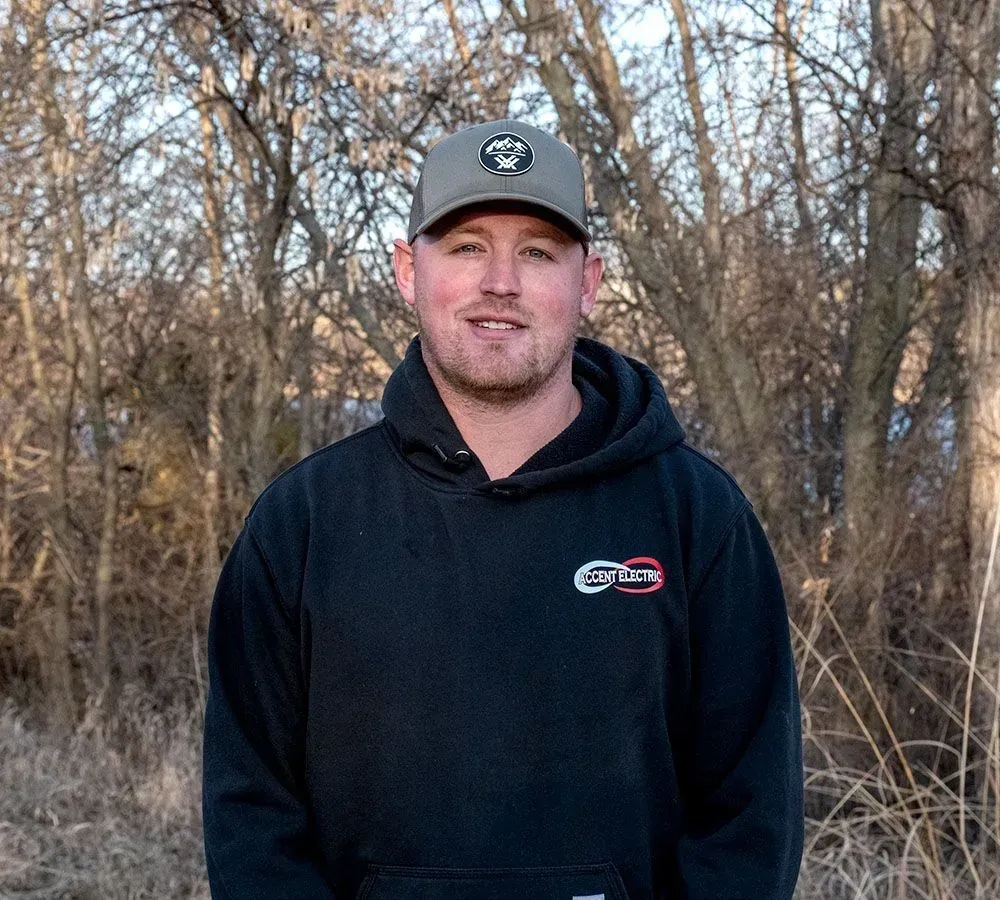 A person wearing a grey baseball cap and a black hooded sweatshirt smiles outdoors in front of bare trees.