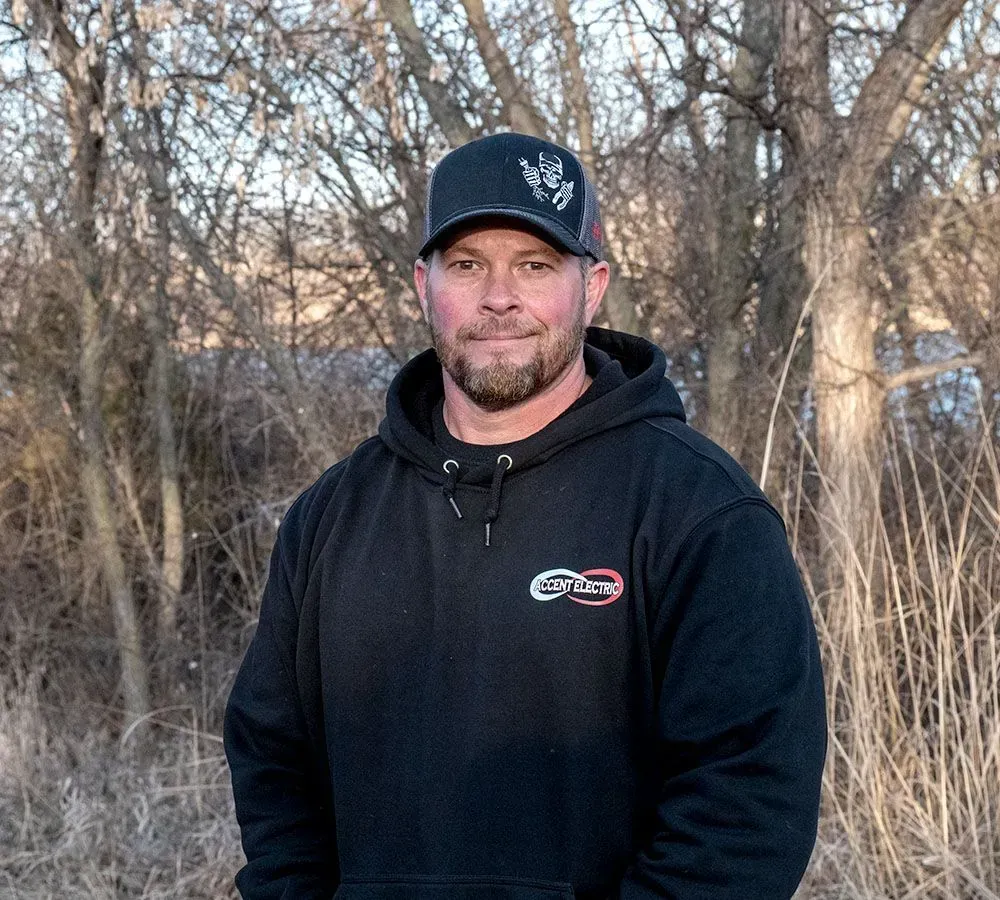 A person wearing a black hoodie and cap stands in a wooded area with bare trees in the background.