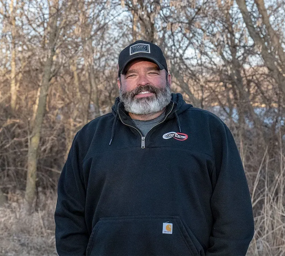 A bearded person wearing a black cap and a black sweatshirt standing in front of leafless trees.