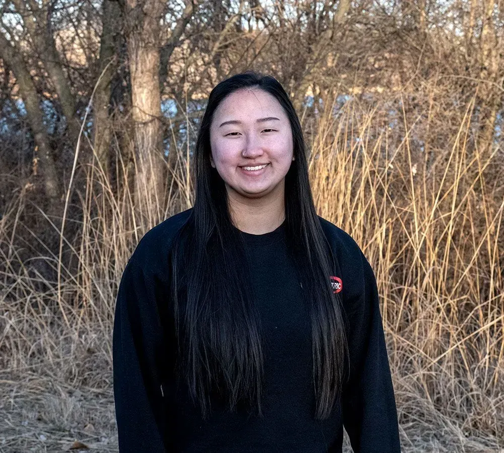 A person with long dark hair smiles while wearing a black sweater, standing in front of dry brush and trees.