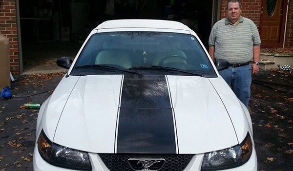 A man is standing in front of a white mustang with black stripes on the hood.
