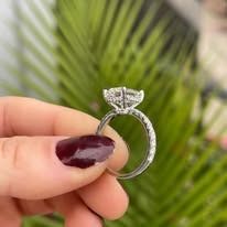 Hand holding a diamond ring with a red nail, green foliage background.