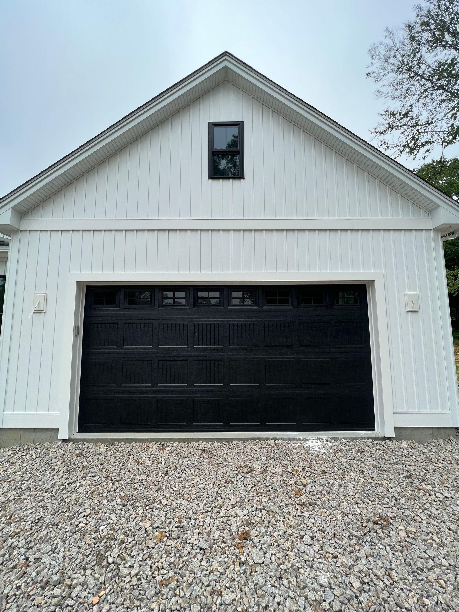 Black garage door on white building with small window above. Gravel driveway.