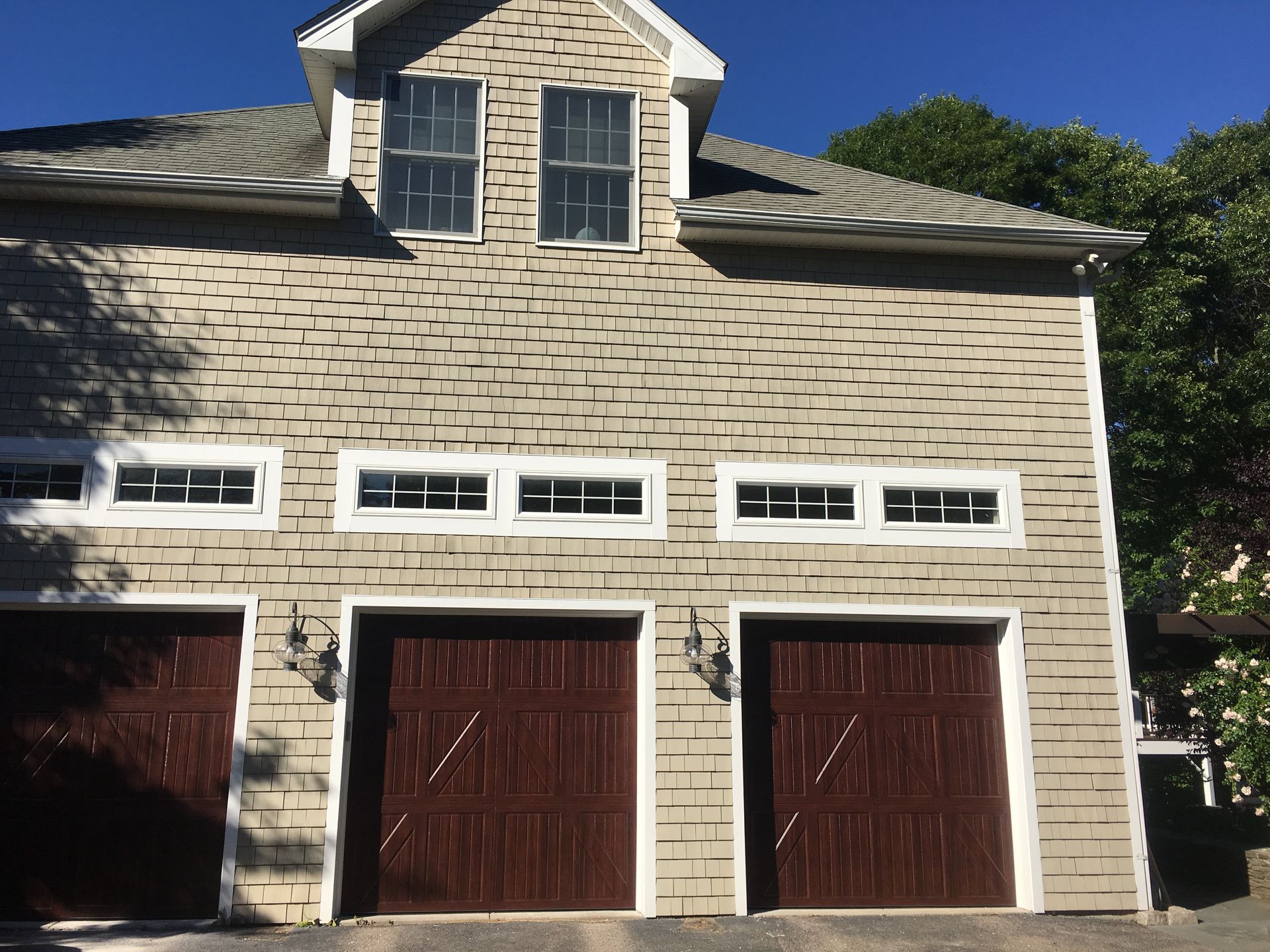 Tan three-car garage with brown doors, windows above doors and in dormer, against a blue sky.