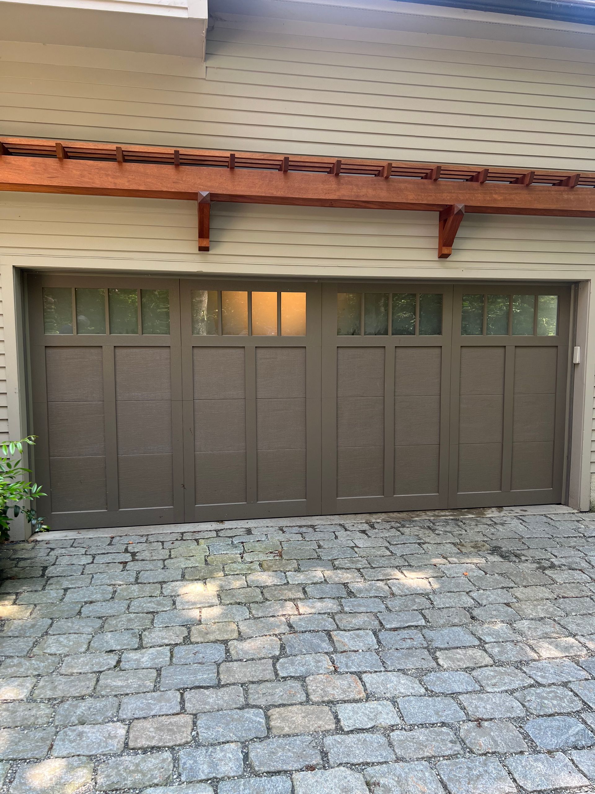 Brown garage door with windows, set in a stone driveway, under a pergola-style roof.