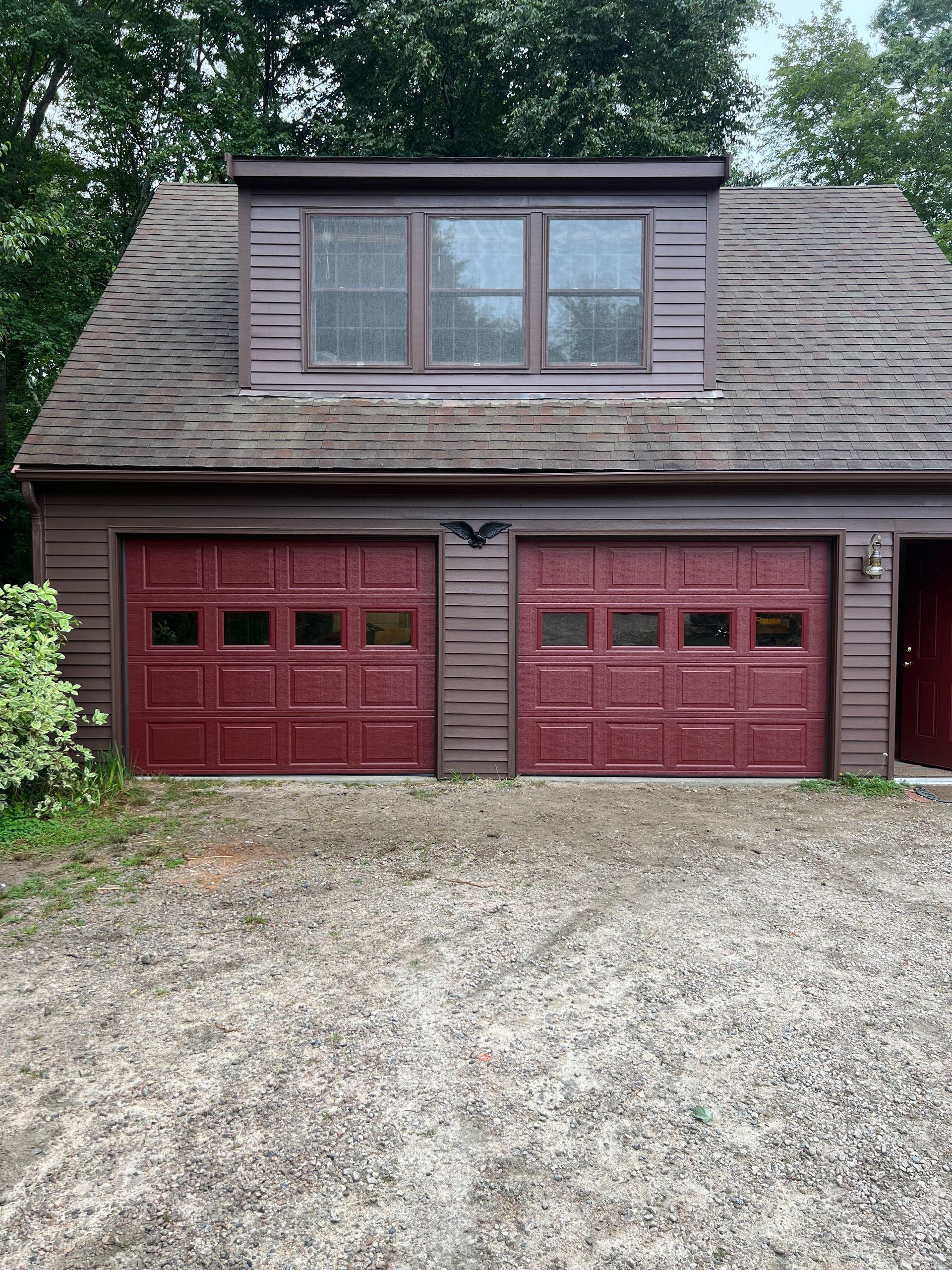 Red garage doors on a brown building with an attic window. Gravel driveway in front.