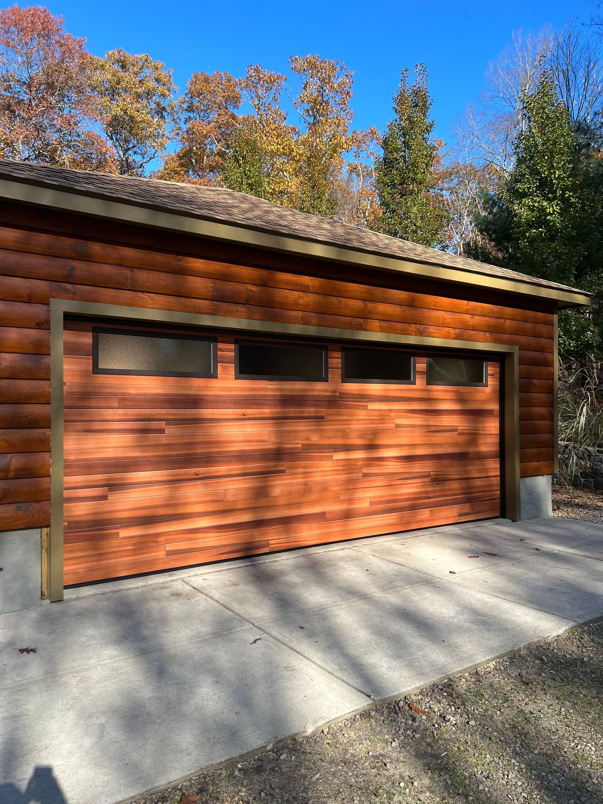 Wooden garage with three small windows above the closed door, set against a backdrop of trees and a blue sky.