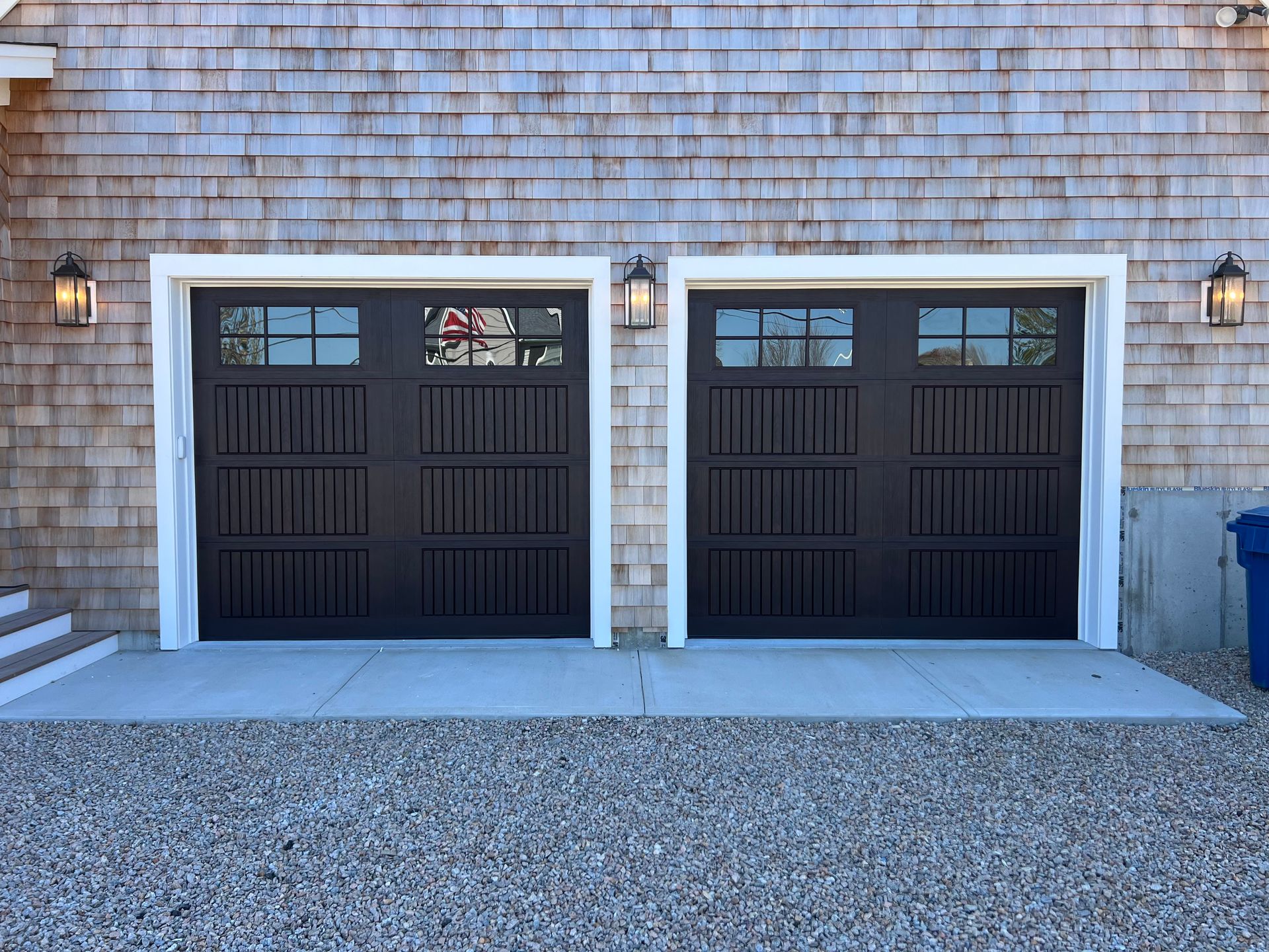 Two black garage doors with white trim and glass panels on a gray concrete pad, gravel driveway, and wood siding.