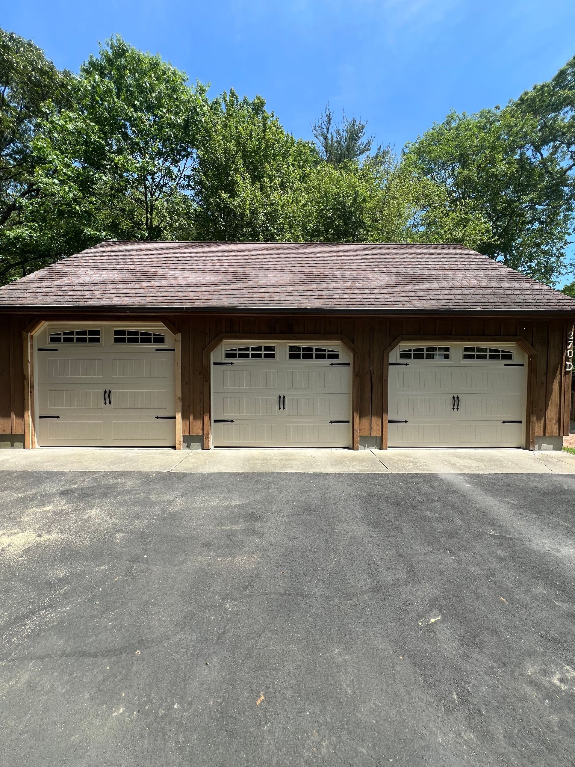 Three-bay garage with white doors, dark brown wooden structure, asphalt driveway, and green trees.
