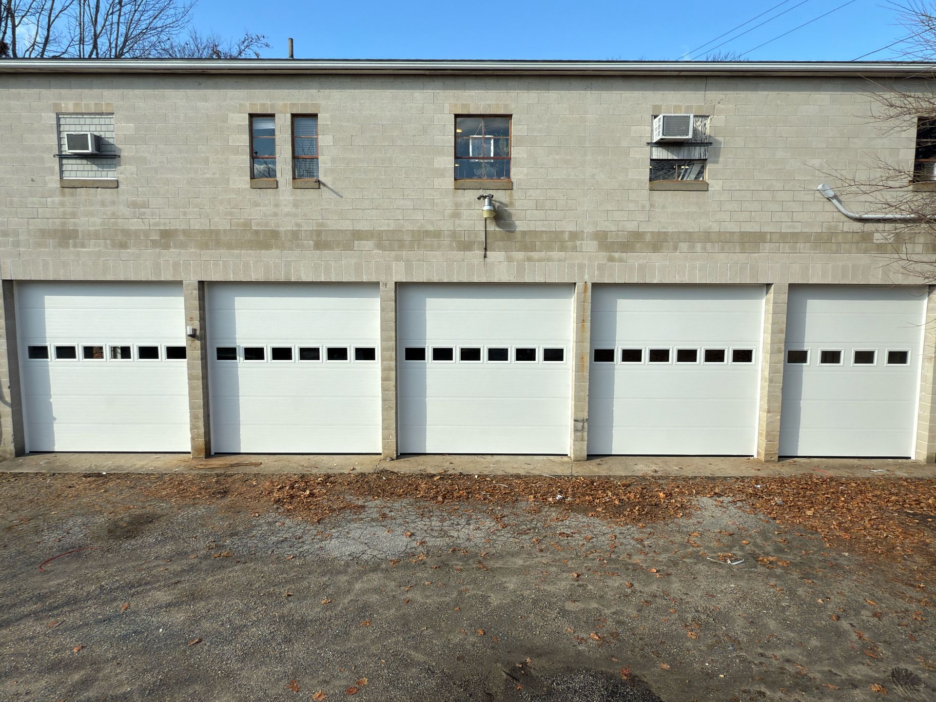 Five white garage doors on a light brick building with small windows and AC units; outdoors on a sunny day.