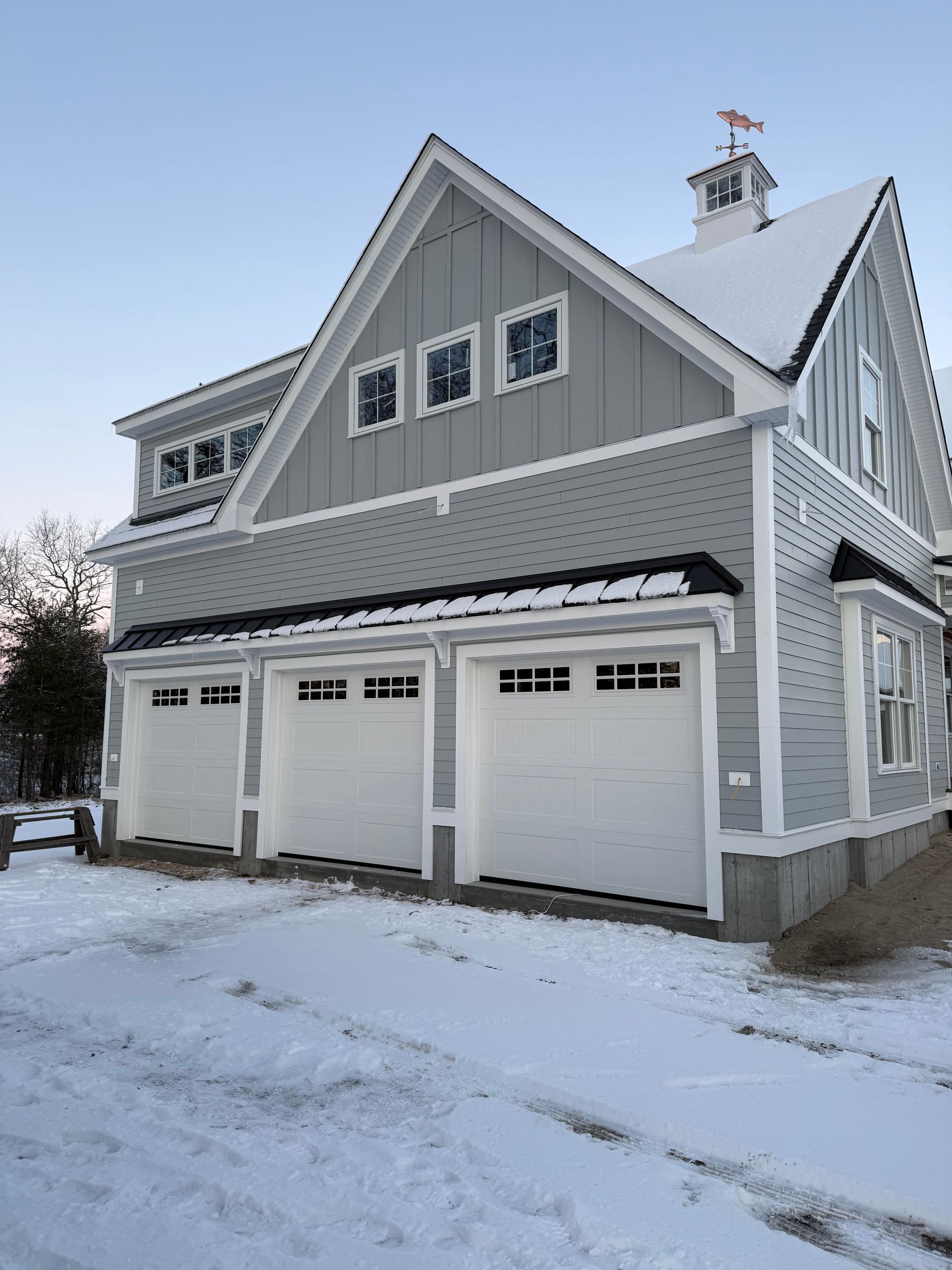 Three-car gray garage with white doors, snow-covered driveway, and a small cupola on roof.