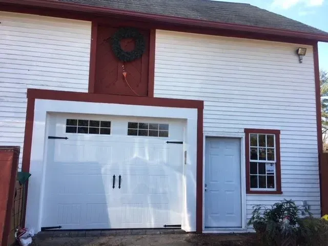 White and red barn-style building with a garage door, side door, and window. A wreath hangs above the garage.