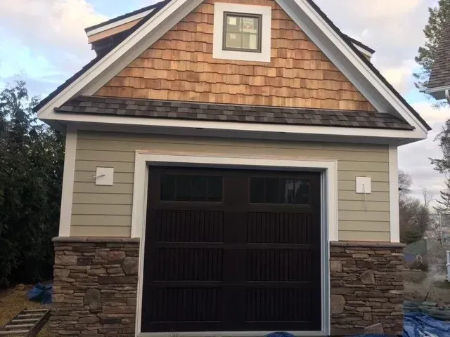 Garage with brown door, tan siding, stone base, and wood shake roof.