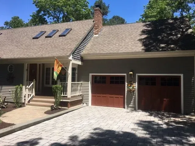 House exterior with brick driveway, brown garage doors, porch, flag, and grey siding.