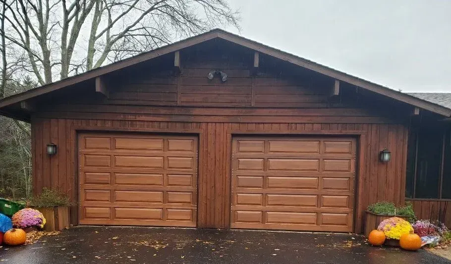 Two-car garage with brown wooden doors and exterior, flanked by fall pumpkins and flowers.