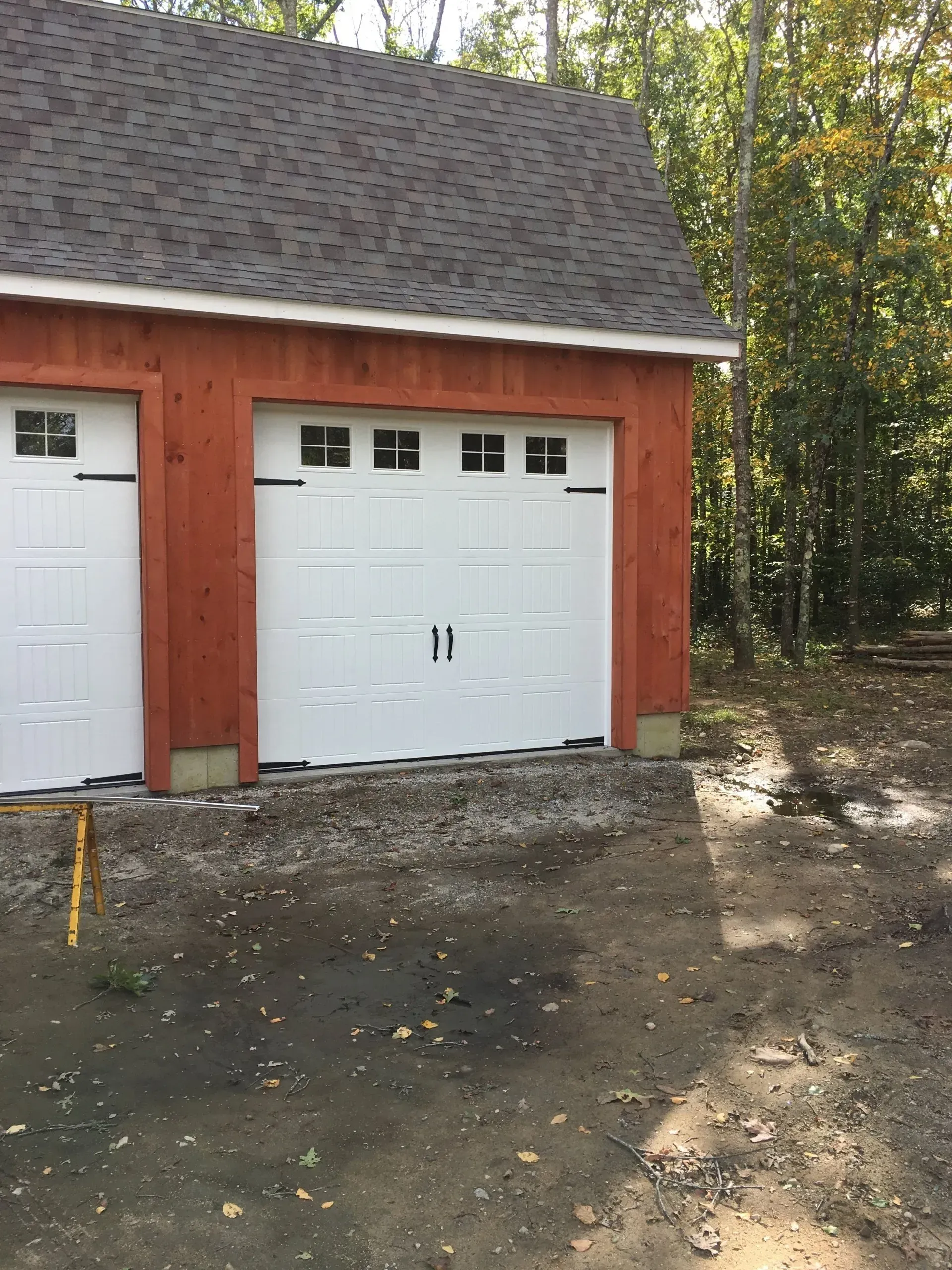 Red-sided garage with white garage doors and a brown roof, set in a wooded area.