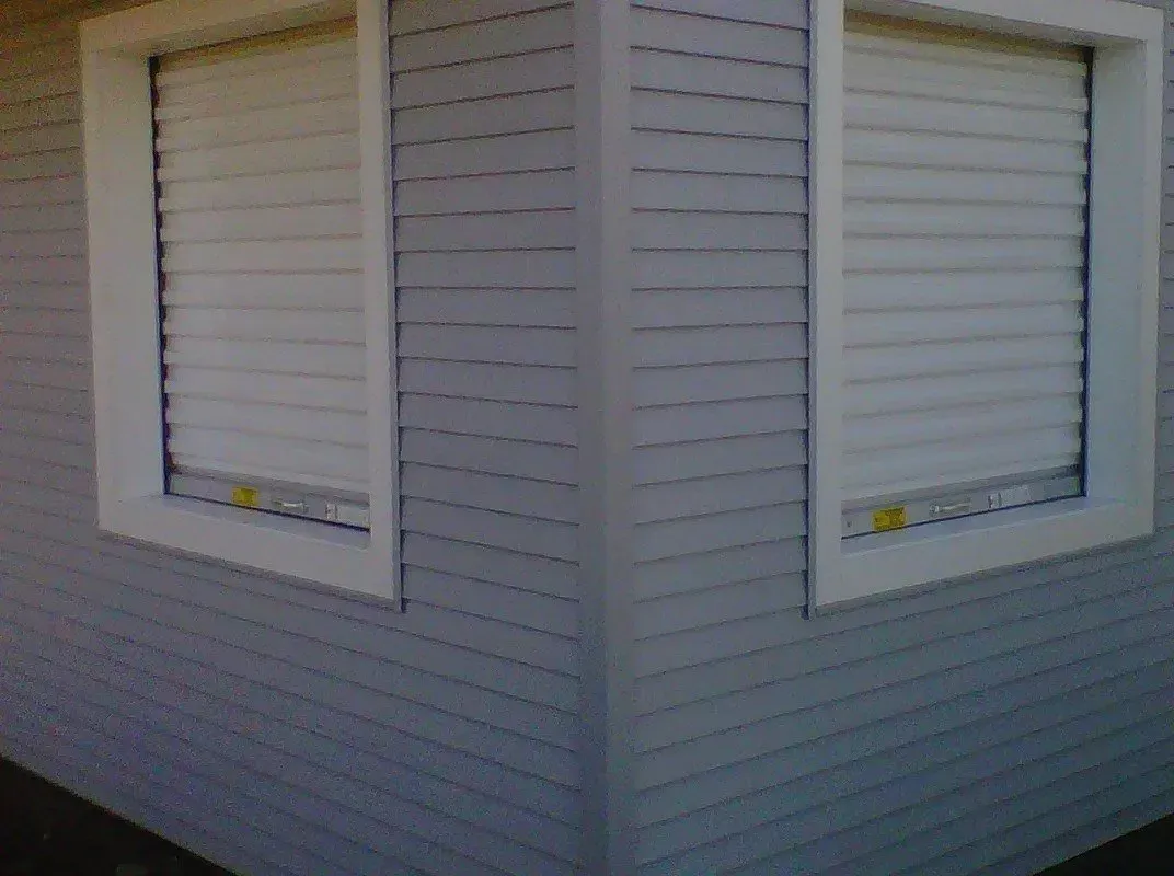 Exterior corner of a building with two windows featuring closed white shutters, gray siding.