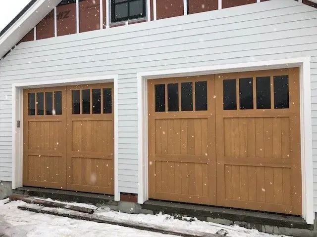 Two tan wooden garage doors with windows on a white house, snow on the ground.