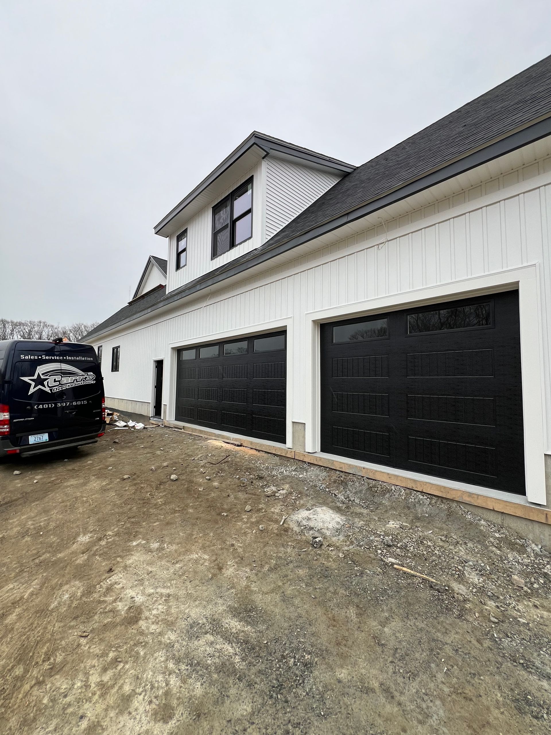 White house with black garage doors, vehicle parked nearby on gravel.