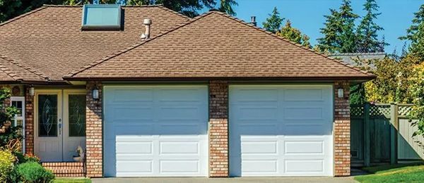 Two-car garage with white doors, brick columns, and brown roof. Green foliage and blue sky in background.