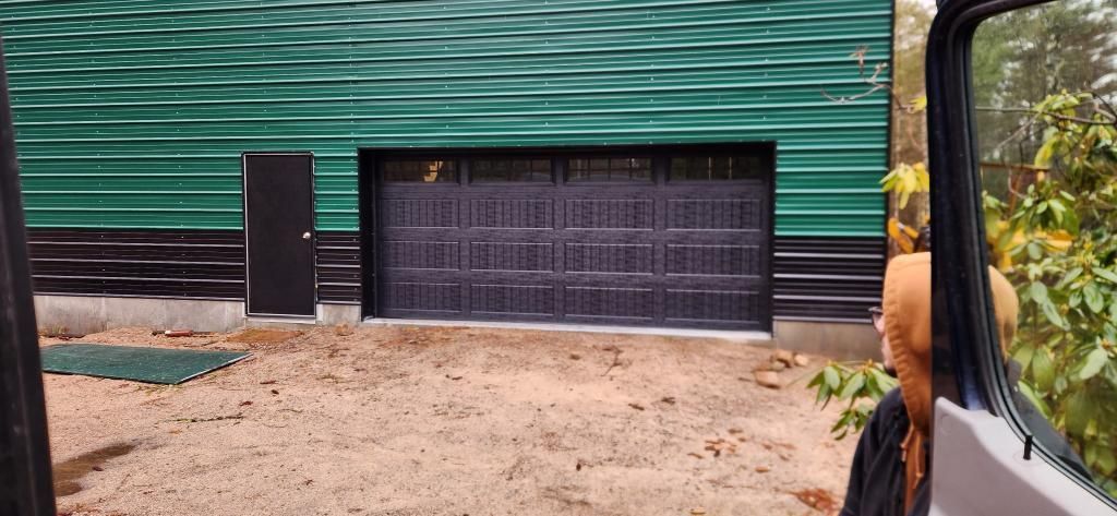 Exterior view of a green metal building with a dark garage door and a side door. A person is in a vehicle looking toward the building.